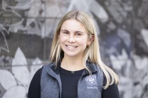 Young woman standing in a light blue grey vest that says "Saint Anselm College Nursing"