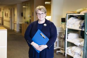 Woman stands in a hospital hallway dressed in a navy blue suit and holding a clipboard