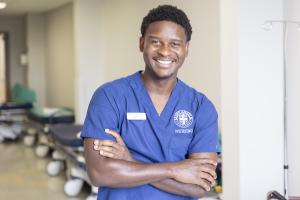 A young man standing in a hospital hallway wearing blue scrubs that say "Saint Anselm College Nursing"