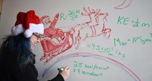 Nicole Gugliucci writing out physics equations on a whiteboard next to a drawing of Santa and his reindeer