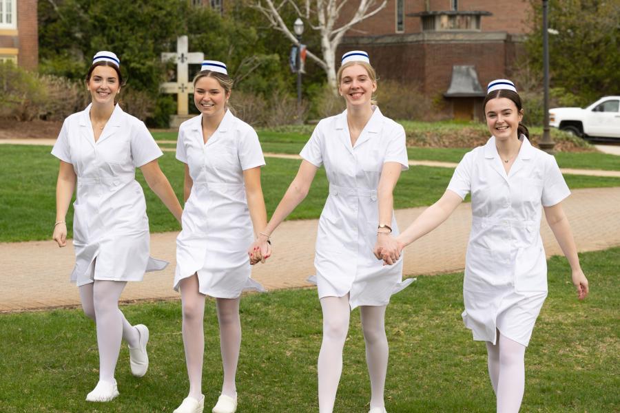 Nurses in pinning ceremony uniforms, holding hands while walking on Alumni Quad